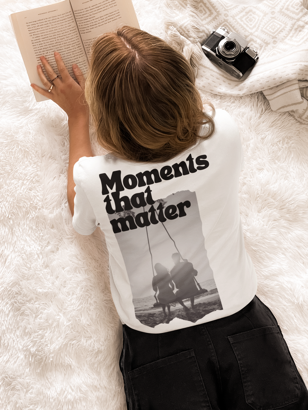 Frau mit braunen Haaren liegt auf weißer Decke, liest Buch, trägt weißes T-Shirt mit schwarzem Aufdruck 'Moments that matter' und Bild zweier Personen auf Schaukel.