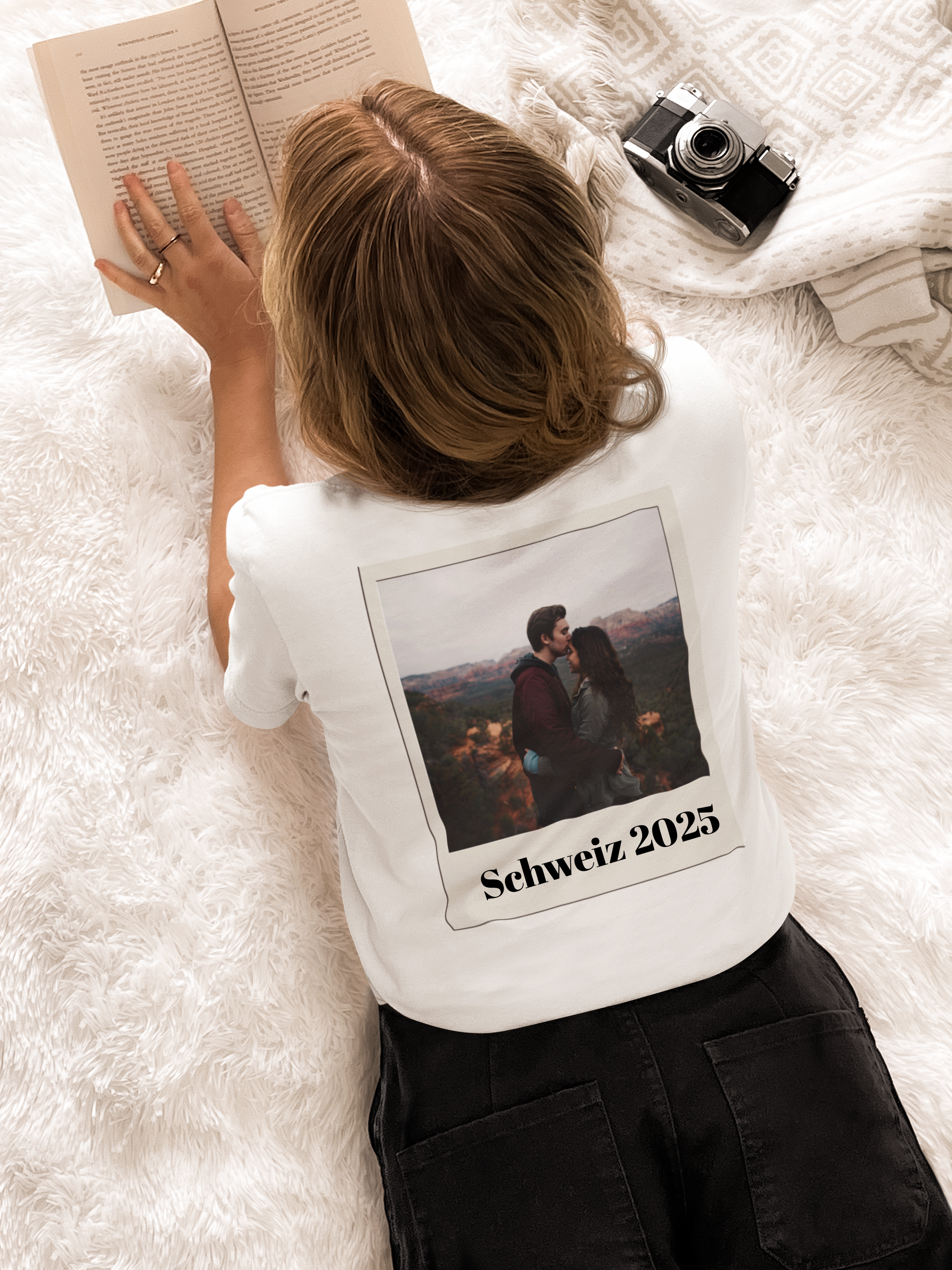Frau liegt auf dem Bauch auf einem weißen Teppich, trägt weißes T-Shirt mit Foto eines Paares und Text 'Schweiz 2025' auf dem Rücken, liest Buch, neben ihr liegt eine Kamera.