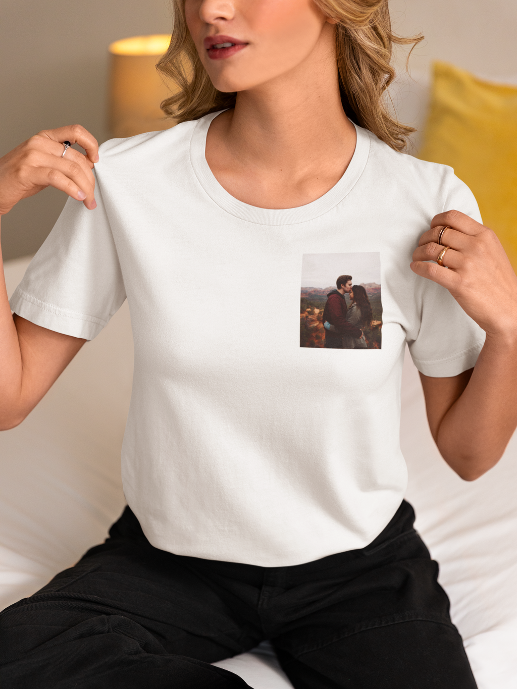 Frau sitzt auf Bett, trägt weißes T-Shirt mit kleinem Foto eines Paares auf der Brust, schwarze Hose, goldene Ringe an den Fingern.