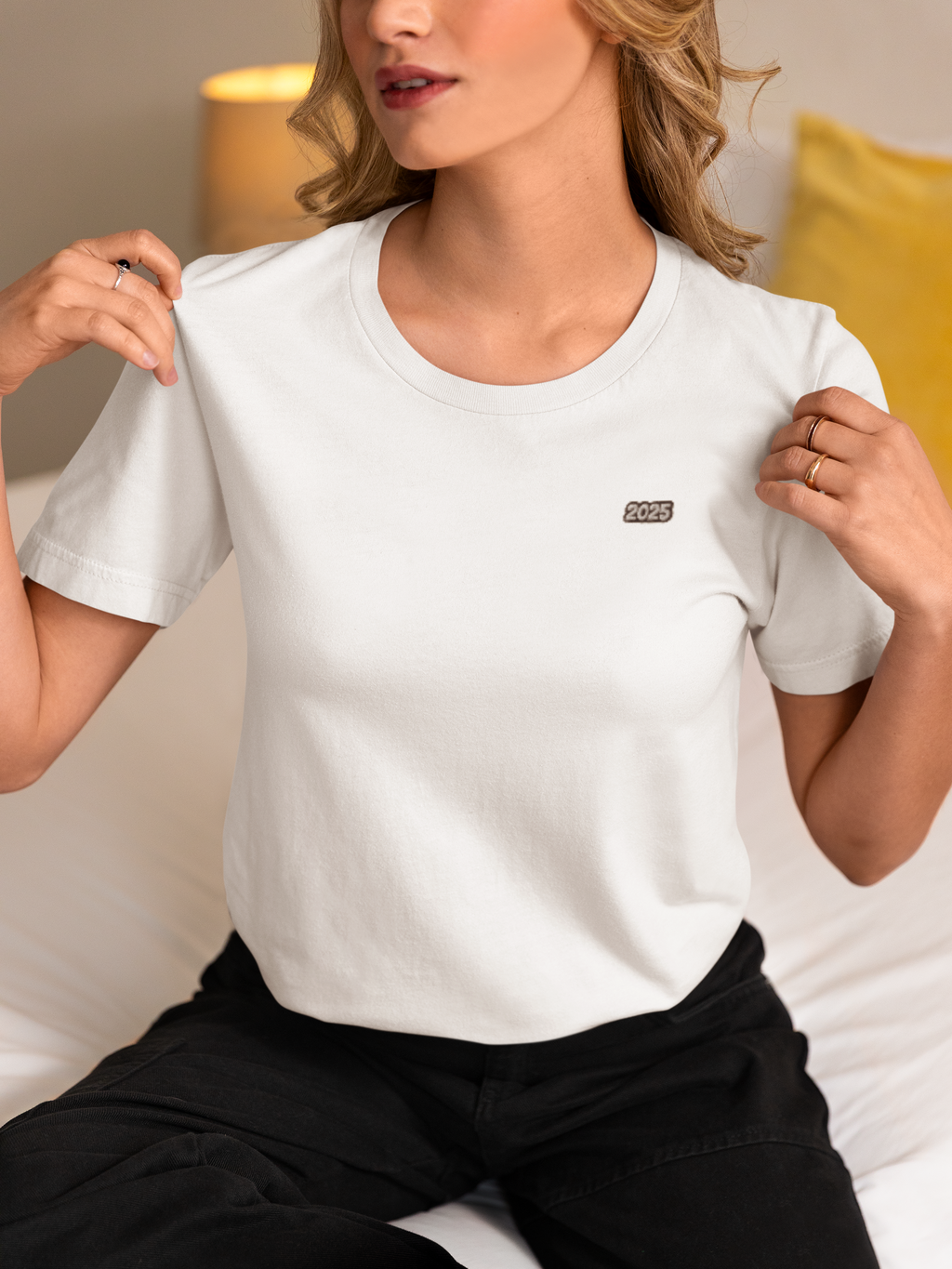 Frau sitzt auf einem Bett, trägt ein weißes T-Shirt mit kleinem Aufdruck '2025' auf der Brust, schwarze Hose, zeigt mit beiden Händen auf das T-Shirt.